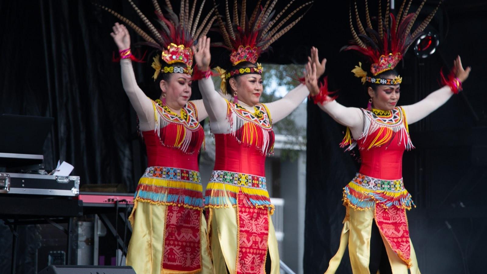 三个女人戴着五颜六色的印尼羽毛头饰跳舞, yellow skirts, 还有串珠腰带和衣领.